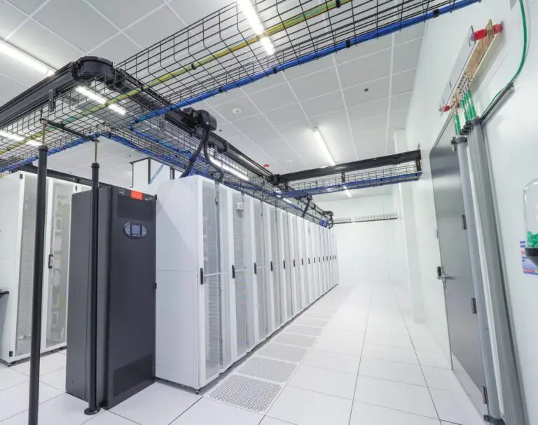 A brightly lit data center room featuring a row of white server cabinets on a raised floor with overhead cable management trays.