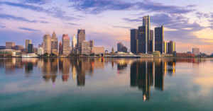 Detroit skyline at sunrise across the Detroit River, with the Renaissance Center and downtown towers reflected in calm water