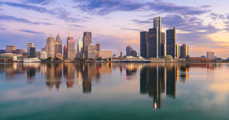 Detroit skyline at sunrise across the Detroit River, with the Renaissance Center and downtown towers reflected in calm water